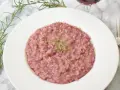 Różowe risotto z prawdziwkami i rozmarynem na białym talerzu, obok widelca i kieliszka wina.
