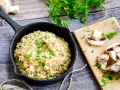 Kremowe risotto z grzybami w czarnej patelni, ozdobione natką pietruszki, obok świeże grzyby na desce.