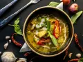 Zielone curry w misce, ozdobione chili i bazylią, otoczone przyprawami.