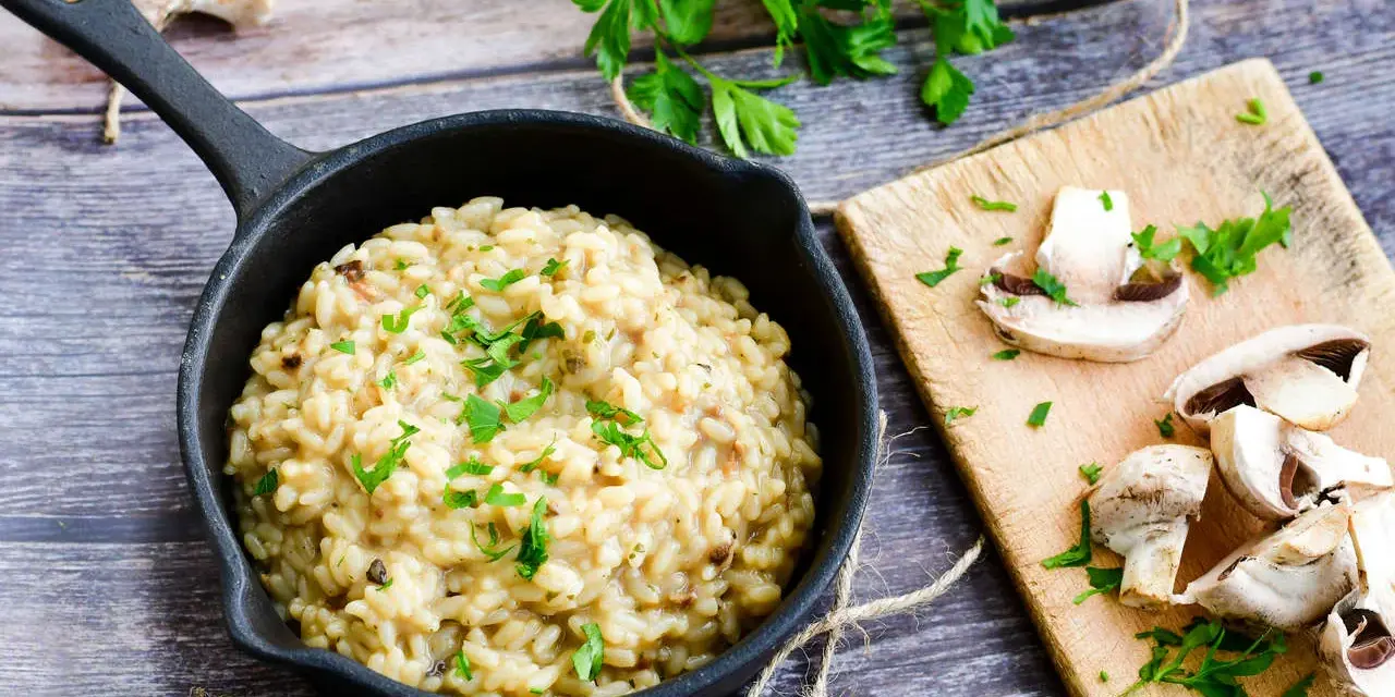Kremowe risotto z grzybami w czarnej patelni, ozdobione natką pietruszki, obok świeże grzyby na desce.