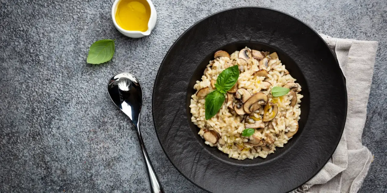 Kremowe risotto z grzybami na czarnym talerzu, obok łyżka i oliwa.