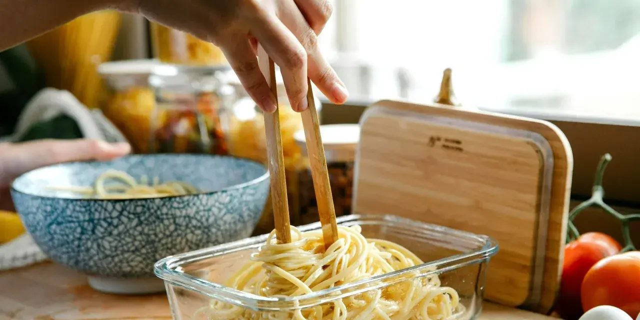 Dłoń trzymająca pałeczki wyciąga makaron spaghetti z szklanego pojemnika.