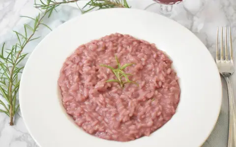 Różowe risotto z prawdziwkami i rozmarynem na białym talerzu, obok widelca i kieliszka wina.