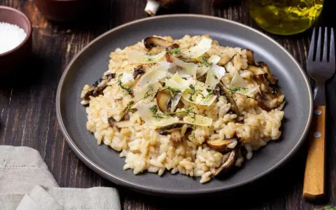 Kremowe risotto z pieczarkami, posypane parmezanem i ziołami, na ciemnym talerzu.