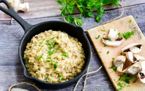 Kremowe risotto z grzybami w czarnej patelni, ozdobione natką pietruszki, obok świeże grzyby na desce.