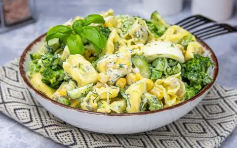 Sałatka z tortellini, brokułów, kukurydzy i jajka w kremowym sosie, udekorowana bazylią.
