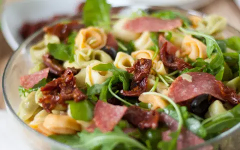 Sałatka z tortellini, suszonymi pomidorami, rukolą i salami w szklanej misce.