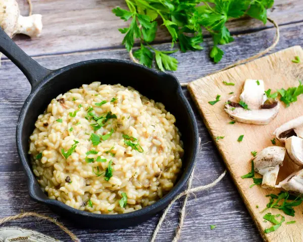 Risotto z grzybami w czarnej patelni, obok deska z pieczarkami i natką pietruszki.