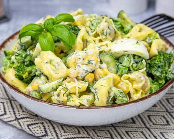 Sałatka z tortellini, brokułów, kukurydzy i jajka w kremowym sosie, udekorowana bazylią.