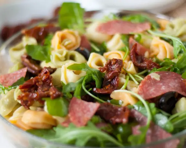 Sałatka z tortellini, suszonymi pomidorami, rukolą i salami w szklanej misce.