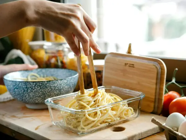 Dłoń trzymająca pałeczki wyciąga makaron spaghetti z szklanego pojemnika.