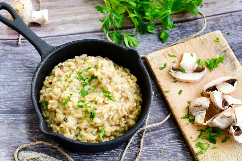 Kremowe risotto z grzybami w czarnej patelni, ozdobione natką pietruszki, obok świeże grzyby na desce.