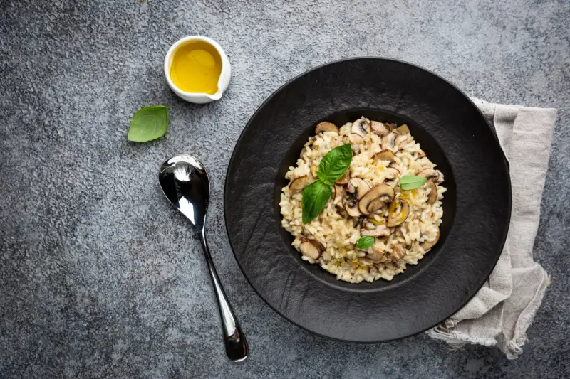 Kremowe risotto z grzybami na czarnym talerzu, obok łyżka i oliwa.